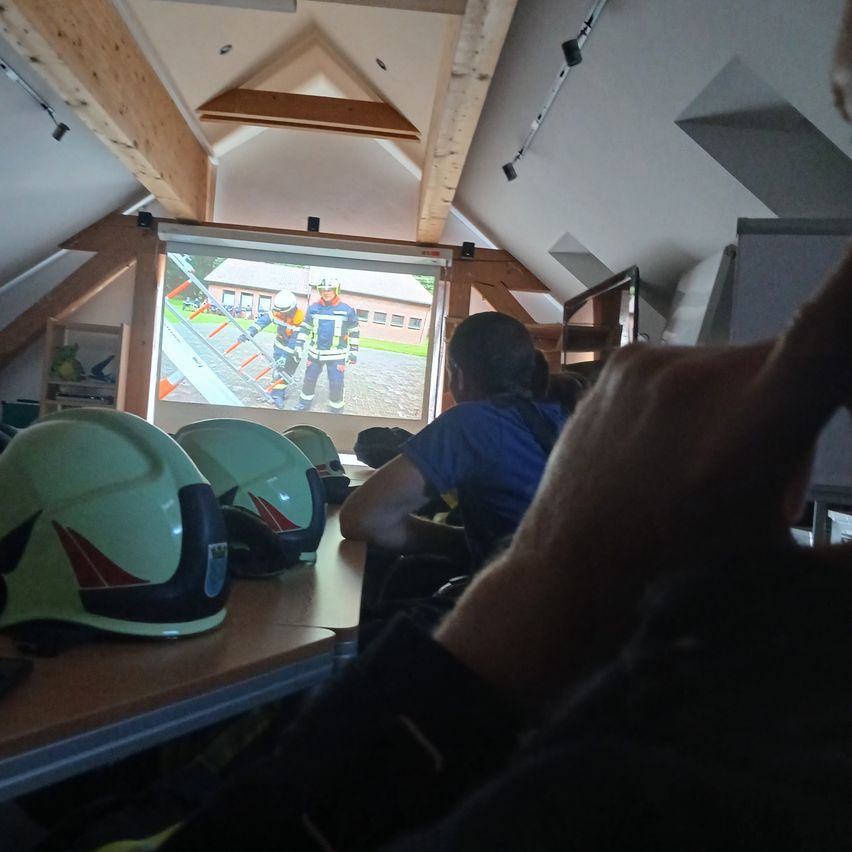 Ein Raum mit Holzbalken an der Decke, Helmen auf einem Schreibtisch und Leuten, die ein Feuerwehrmann-Video auf einer Projektionsleinwand ansehen.