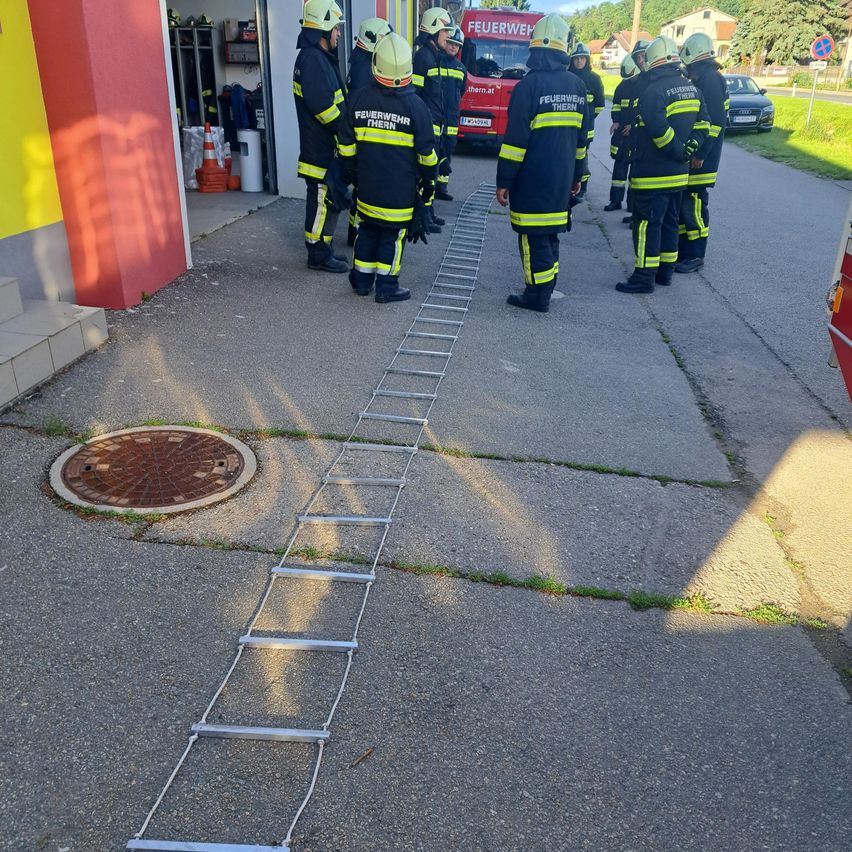 Feuerwehrleute stehen in einer Reihe, tragen Helme und Uniformen, mit einem roten Feuerwehrwagen, der an einer Feuerwehrstation geparkt ist. Eine Leiter liegt vor ihnen auf dem Boden.