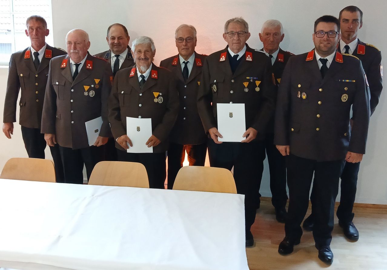 Sieben Männer in Militäruniformen stehen in einem Raum und halten Urkunden. Sie lächeln, und ein Tisch mit einem weißen Tuch steht vor ihnen.