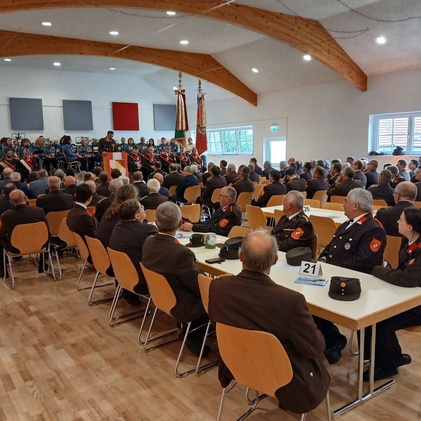 Eine große Gruppe von Menschen in Militäruniformen und Hüten sitzt in einem Saal. Vorne spielt eine Band, und es gibt Fahnen auf Stangen. Der Saal hat Holzböden und Balken.