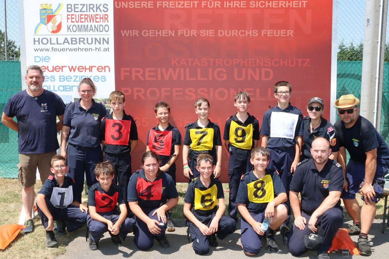 A group of young firefighters, wearing uniforms with numbers, pose for a photo in front of a red sign.