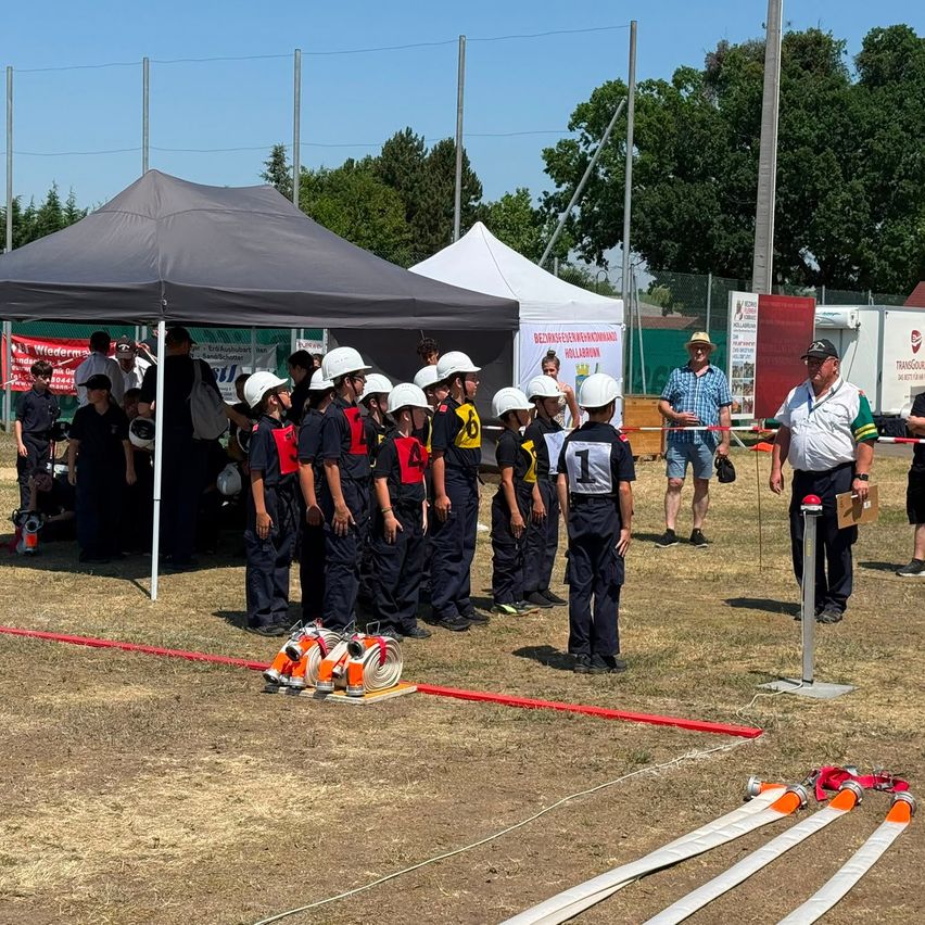 A group of children in uniforms and helmets gather under a tent. A man holds a board with a target. Fire extinguishers are on the ground.
