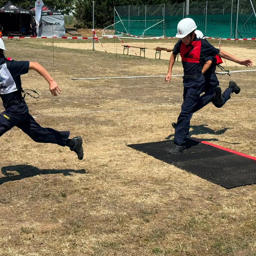 Two individuals in protective gear sprint on a field. One wears a white helmet, while the other wears a red and blue vest. Both are in the midst of a race.
