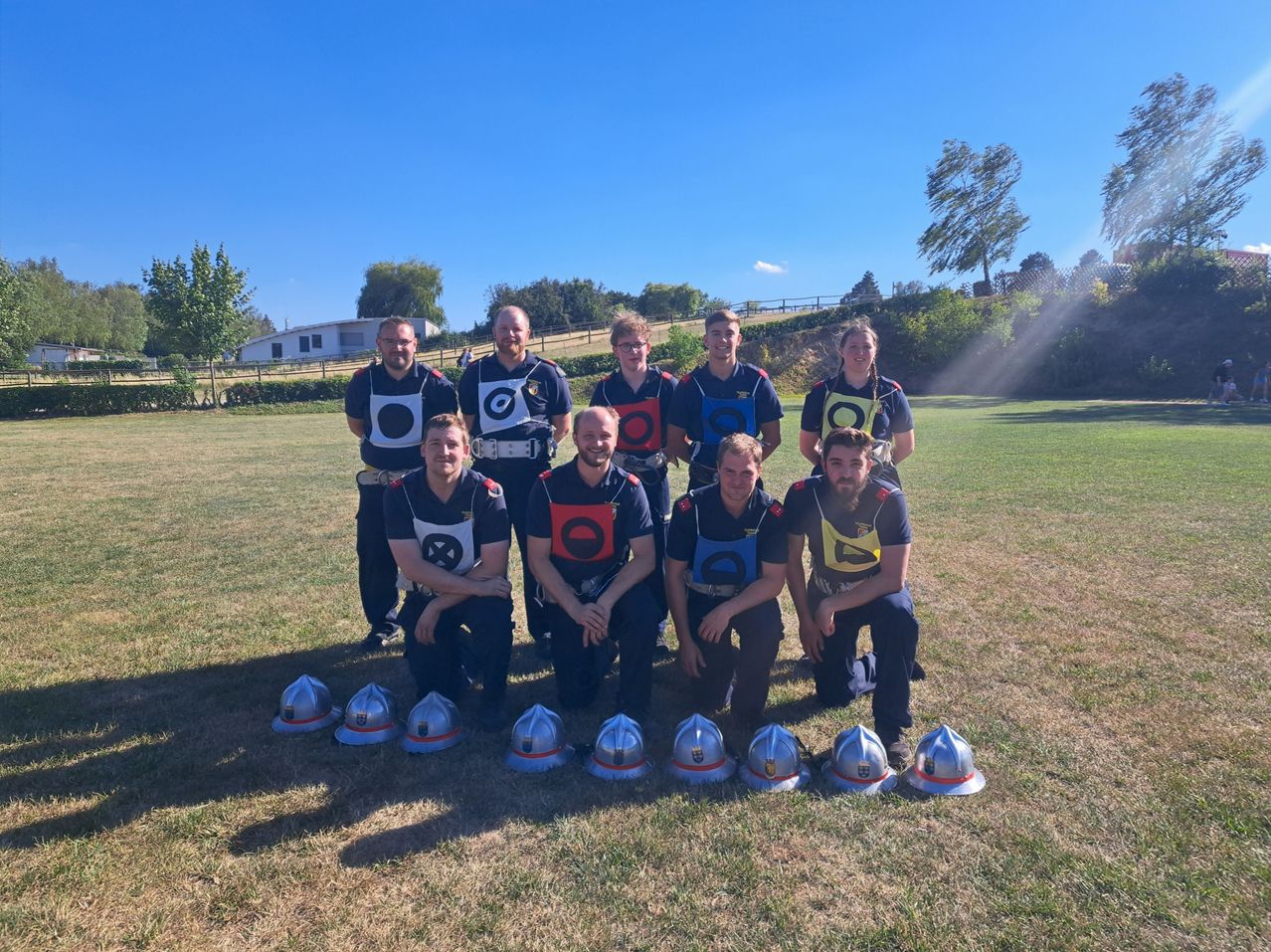 A group of individuals dressed in matching uniforms poses for a photo on a grassy field, with helmets arranged in front of them and trees in the background.