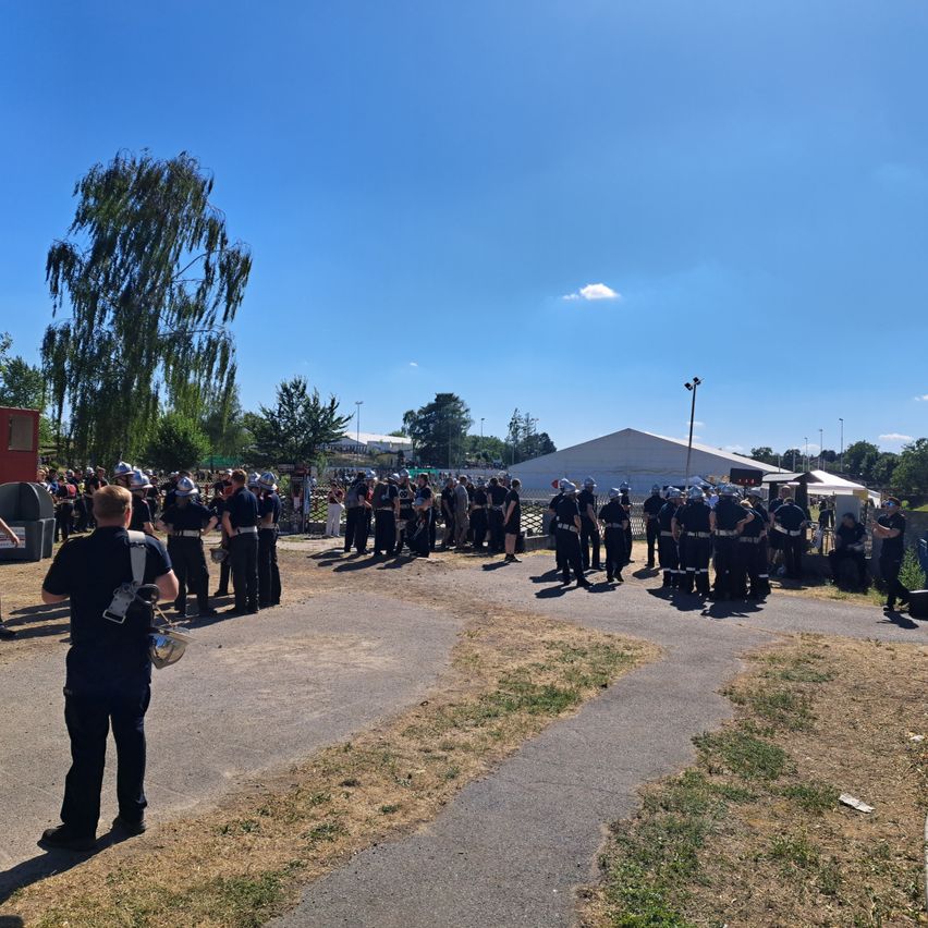 A large group of people in uniform are gathered in an open area, possibly a training exercise. They are standing in rows with helmets on, and one person is holding a shield. In the distance, there are tents and streetlights.