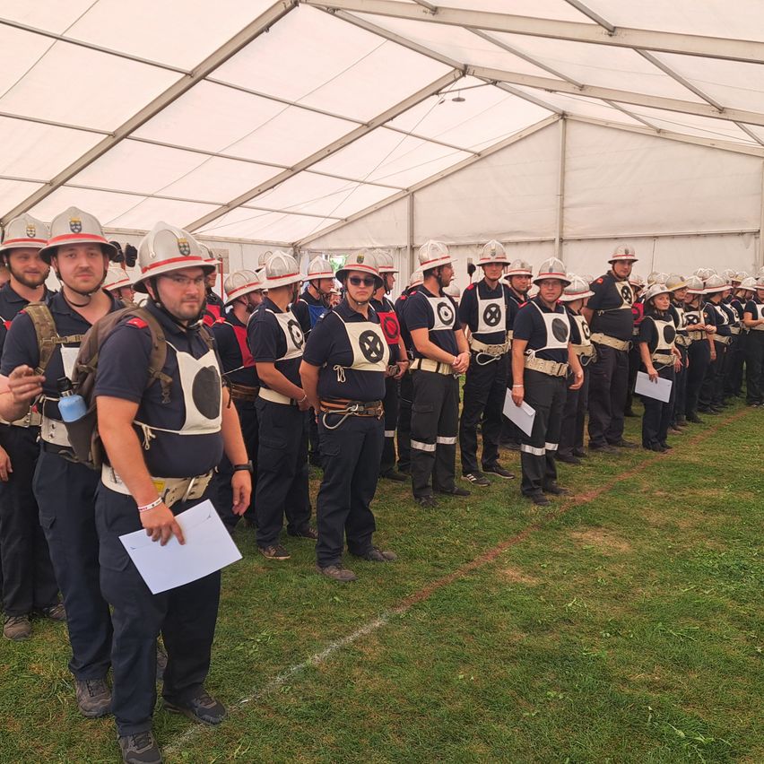 A group of firefighters in uniform, equipped with helmets, stand in line under a white tent on a grassy field. They are holding papers.