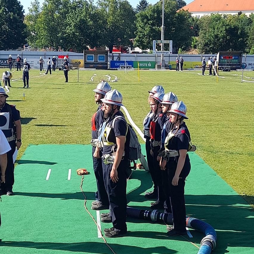 A group of firefighters, wearing helmets and safety gear, stands on a green field with a hose and equipment. In the background, a building and trees are visible.