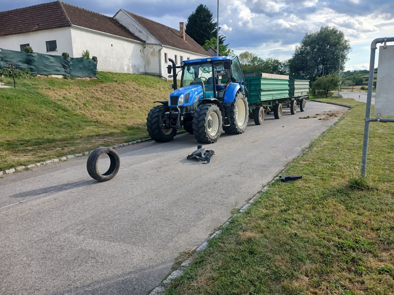 A blue tractor is parked on a road with a house and greenery behind it. The tractor has two green trailers attached. A tire is on the road, and other objects are nearby.