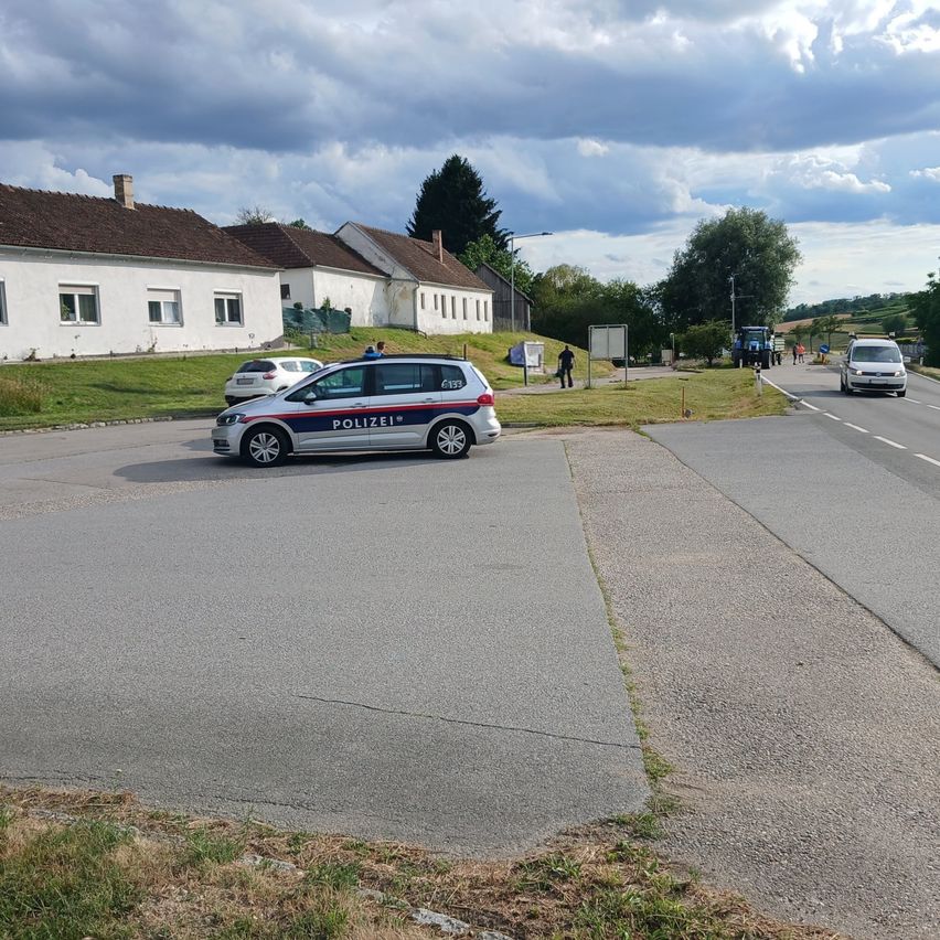 A police car is parked in a small town. In the background, there are several buildings and a tractor. People are standing on the roadside.
