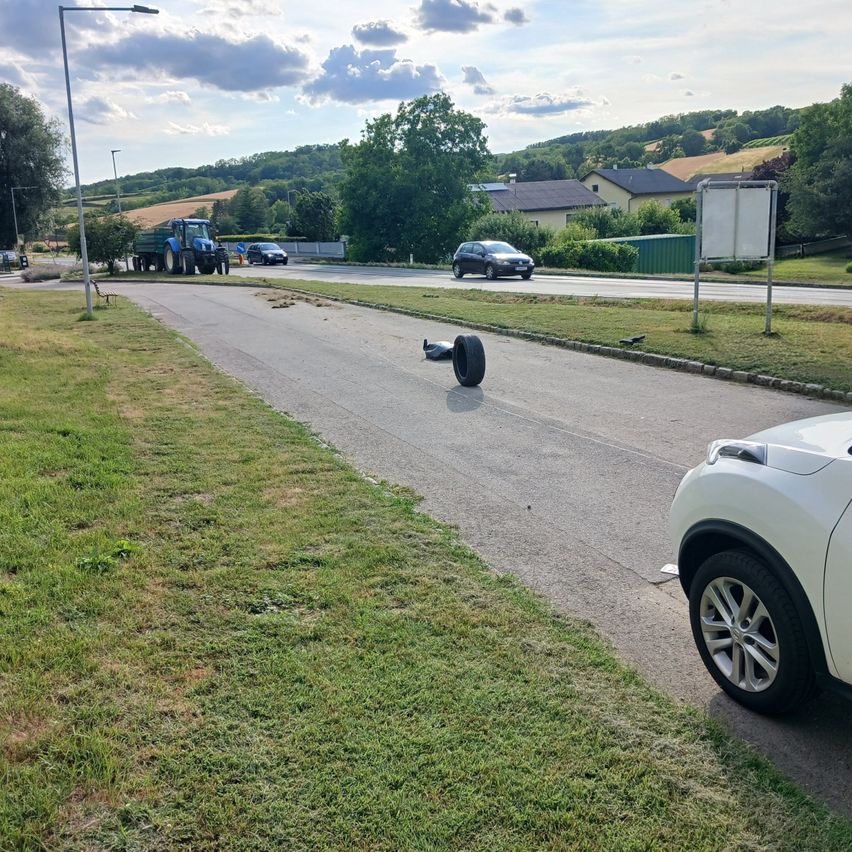 A white car is parked on the side of the road. A tire is on the road, and a blue truck is parked nearby.