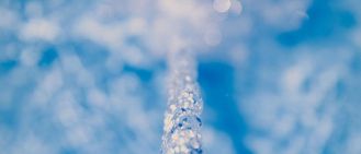 Eine Nahaufnahme eines Wassertropfens, der von einem Wasserhahn in einen Pool fällt, dabei Wellen schlägt und unter dem Licht funkelt.