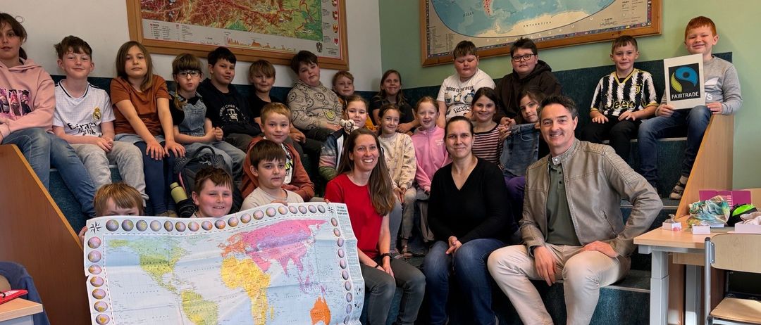 Eine Gruppe von Kindern und Erwachsenen in einem Klassenzimmer, einige sitzen auf einer Bank und andere stehen. Eine Frau hält eine große Weltkarte, und es liegen Papiere und ein Stift auf dem Boden. Zwei große Karten und ein Schild hängen an den Wänden.