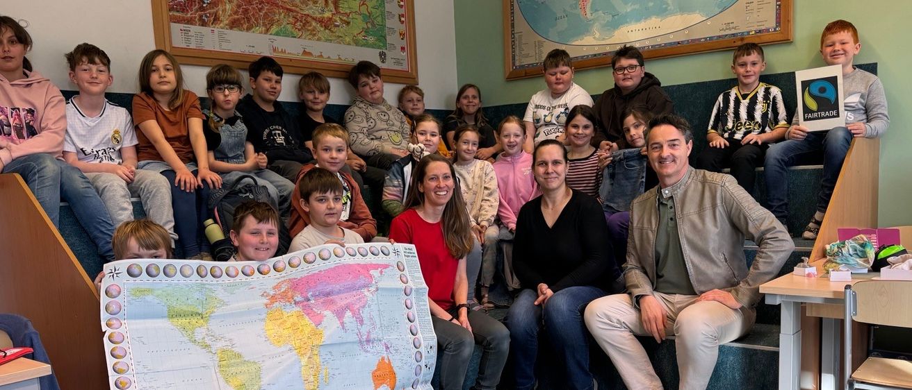 Eine Gruppe von Kindern und Erwachsenen in einem Klassenzimmer, einige sitzen auf einer Bank und andere stehen. Eine Frau hält eine große Weltkarte, und es liegen Papiere und ein Stift auf dem Boden. Zwei große Karten und ein Schild hängen an den Wänden.