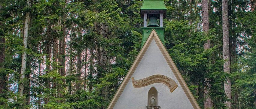Eine kleine Kapelle mit einem grünen Turm steht in einer Waldlichtung, umgeben von hohen Bäumen. Sie verfügt über eine Tür mit Buntglas und eine Nische mit einer Statue. In der Nähe steht eine kleine Bank.