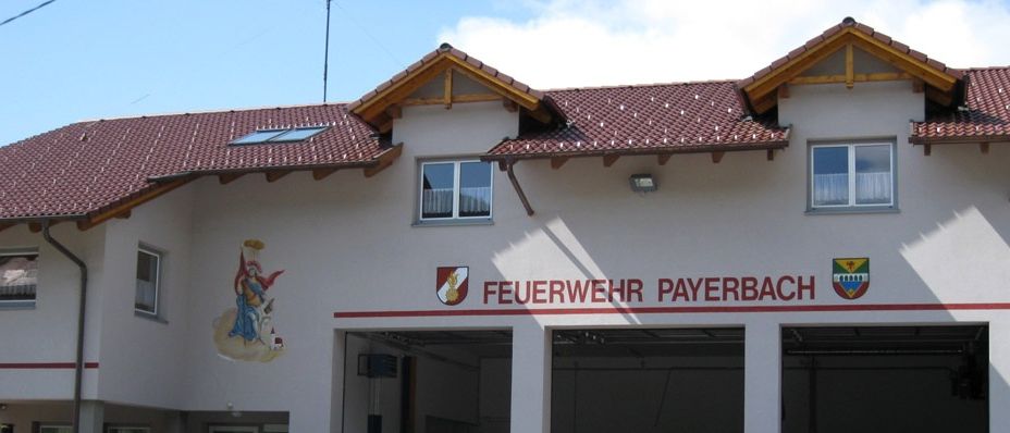 A marching band stands in front of the Feuerwehr Payerbach building, with people gathered in a large assembly. The building has a red roof, windows, and an emblem above the entrance.