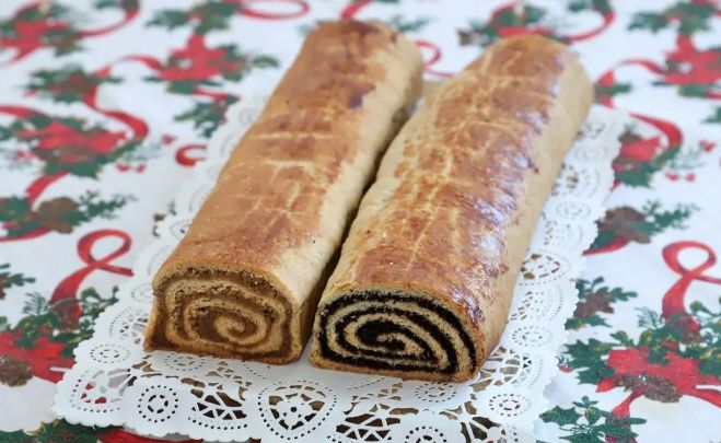 Zwei Schokoladenrollen sind auf einer weißen Spitzenunterlage auf einem weihnachtlich gestalteten Tischtuch ausgestellt.