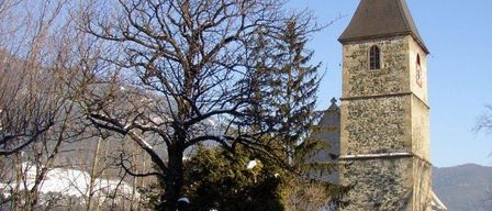 Eine Kirche mit Turm und Uhr steht in einer verschneiten Landschaft. Eine Bank ist in der Nähe, und Bäume umgeben das Gebiet. Berge sind in der Ferne sichtbar.