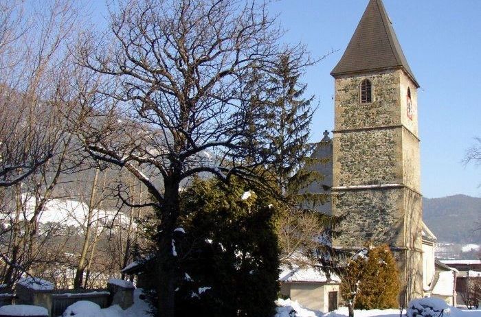 Eine Kirche mit einem hohen Turm steht im Schnee. Sie hat eine Uhr und ist von Bäumen umgeben. Der Himmel ist klar und blau.