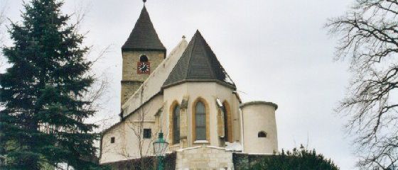 Eine Steinkirche mit Glockenturm steht in einer verschneiten Landschaft. Bäume umgeben die Kirche, und ein grüner Laternenpfahl steht davor.