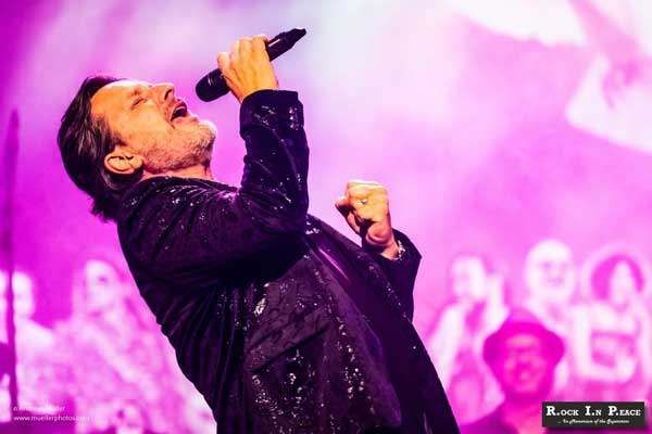 Ein Mann singt in ein Mikrofon vor einem lila Hintergrund. Er trägt einen schwarzen Anzug und einen Ring am Finger. Hinter ihm sind Menschen verschwommen. Das Bild hat einen Wasserzeichen mit dem Text Rock In Peace.