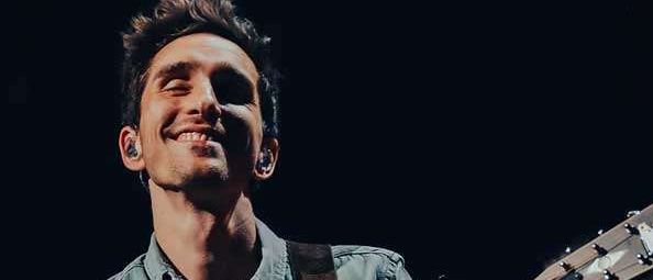 Ein lächelnder Mann in einem grünen Hemd spielt auf der Bühne eine akustische Gitarre, mit einer Frau hinter ihm.