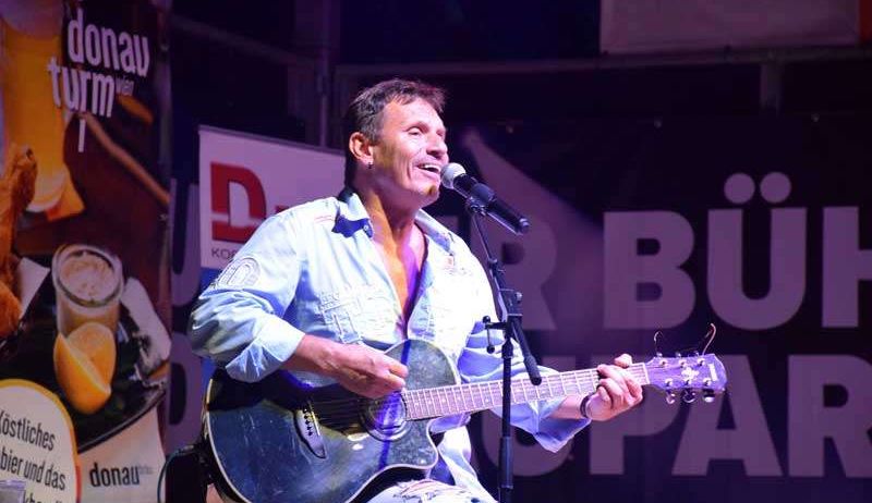 Ein Mann spielt Gitarre und singt auf der Bühne vor einem Mikrofon. Dahinter steht ein Banner mit der Aufschrift 'R Buer'. Links neben ihm steht ein Tisch mit einem Teller und einer Zitronenscheibe.