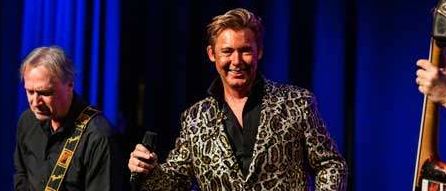 Drei Männer spielen auf der Bühne, einer mit einem Mikrofon, ein anderer mit einer Gitarre und der dritte mit einem Bass, beleuchtet von Scheinwerfern.