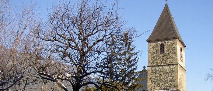 A church with a steeple, bell tower, and clock stands in a snowy landscape. Trees surround the church, and a bench is visible on the left.