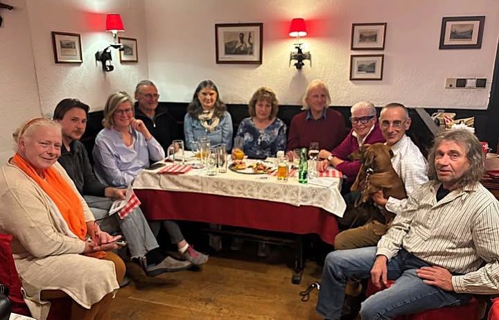 Eine Gruppe von Menschen sitzt um einen Tisch mit Essen, Getränken und einem Hund in einer gemütlichen Restaurantatmosphäre. Gerahmte Bilder an der Wand und rote Lampen tragen zur Stimmung bei.