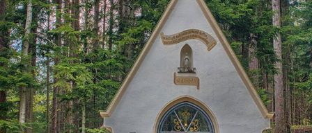 A small chapel with a green steeple is surrounded by tall trees. The chapel has a stained glass window and a bell. A bench is placed in front.