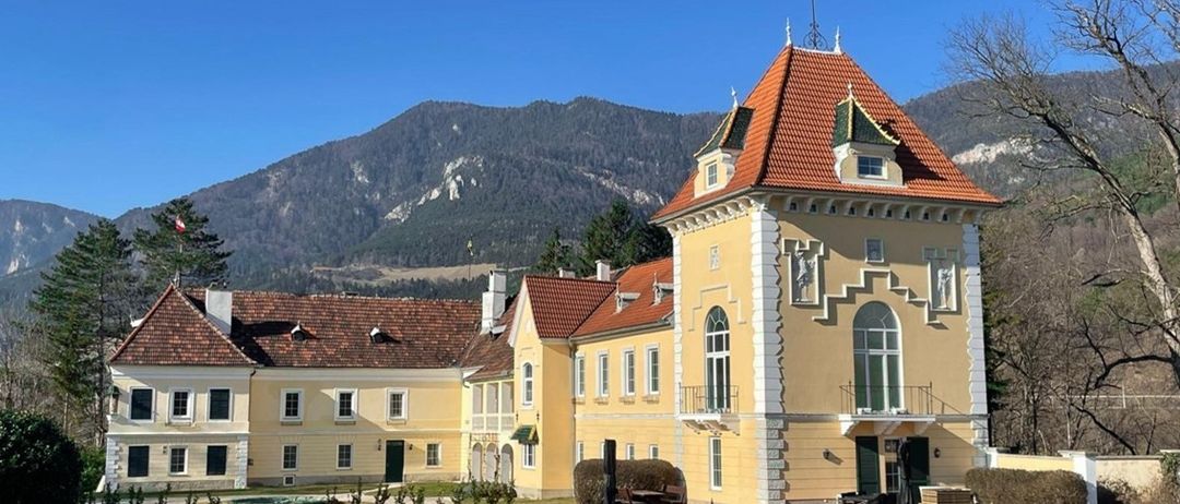 Ein gelbes Gebäude mit rotem Dach und Turm steht auf einem Feld. Im Hintergrund befinden sich Berge unter einem klaren blauen Himmel.