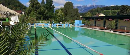 An outdoor pool with blue and green lanes, surrounded by lush trees and mountains. The water is clear and clean, with a white and red lane rope.