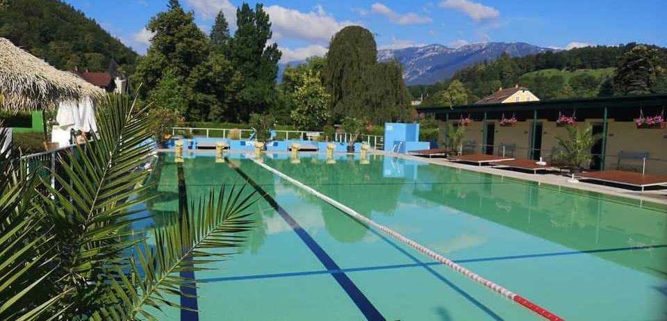 Ein Freibad mit blauen und grünen Bahnen, umgeben von üppigem Baumbestand und Bergen. Das Wasser ist klar und sauber, mit einem weißen und roten Bahnseil.
