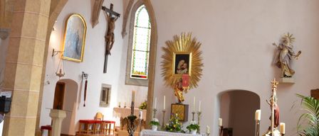 Das Innere einer Kirche mit Bögen, einer goldenen Sonne und einem Kruzifix. Kerzen und Blumen schmücken den Altar, während Statuen und Gemälde die Wände zieren.