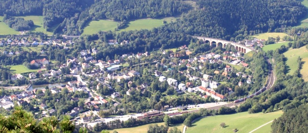 Eine Luftaufnahme einer Stadt mit vielen Gebäuden, einem Fluss und einer Brücke. Die Stadt ist von grünen Feldern und Bäumen umgeben.