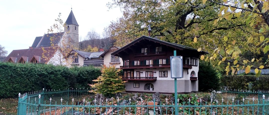 Ein kleines Haus mit Balkonen ist von einem grünen Zaun umgeben. Bäume und Büsche sind im Vordergrund. Ein Kirchturm ist in der Ferne sichtbar.