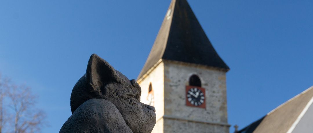 Eine Steinfigur eines Gargoyles ist auf einem Sims vor einem Kirchturm mit einer Uhr angebracht. Der Gargoyle schaut nach oben, und der Himmel ist klar und blau.