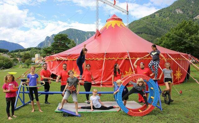Eine Gruppe von Kindern und Erwachsenen übt Zirkustricks vor einem roten Zelt mit Bergen im Hintergrund.