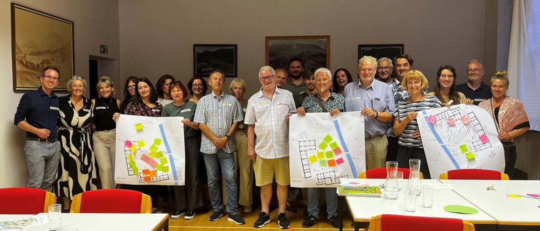 Eine Gruppe von Menschen in einem Raum, die große Papiere mit Grundrissen und Notizen hochhalten und für ein Foto lächeln.