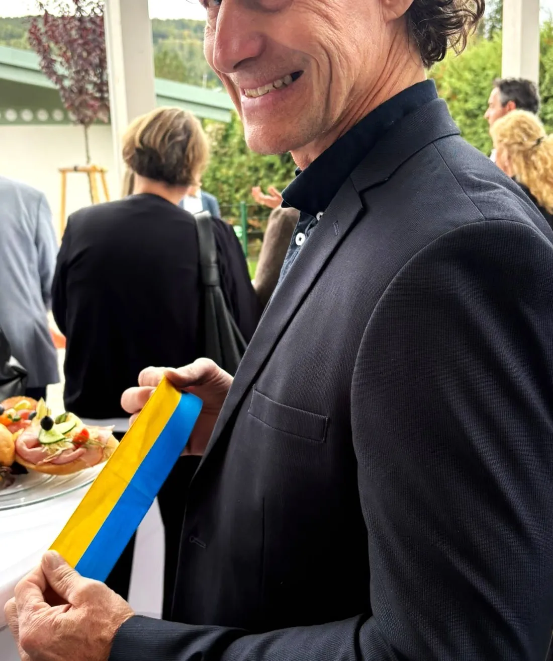 Ein Mann in einem schwarzen Anzug hält ein blaues und gelbes Band und lächelt in die Kamera. Hinter ihm steht ein Tisch mit Essen und Menschen in einem Garten.