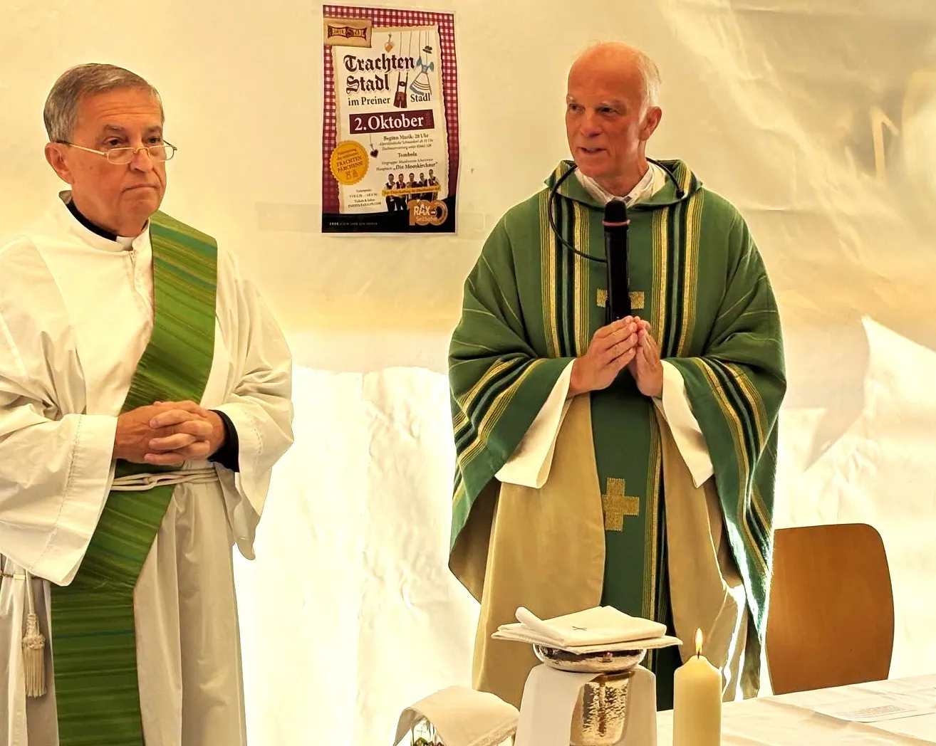 Zwei Priester stehen vor einem Tisch mit einer Kerze und Blumen. Ein Priester hält ein Mikrofon und scheint zu sprechen.