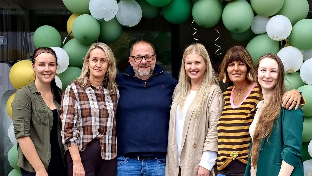 Eine Gruppe von sechs Menschen steht zusammen und lächelt für ein Foto. Sie stehen vor einem Hintergrund aus grünen und weißen Luftballons.