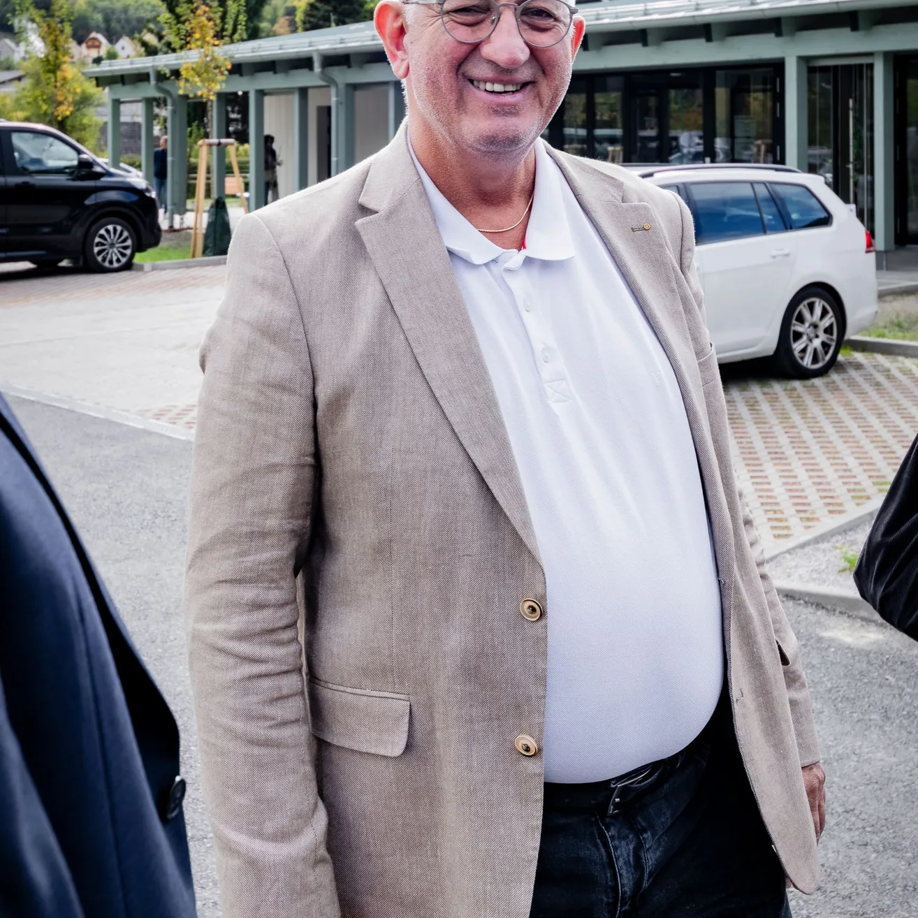 Ein älterer Mann in einem weißen Hemd und einem hellbraunen Blazer lächelt und steht vor einem Gebäude.