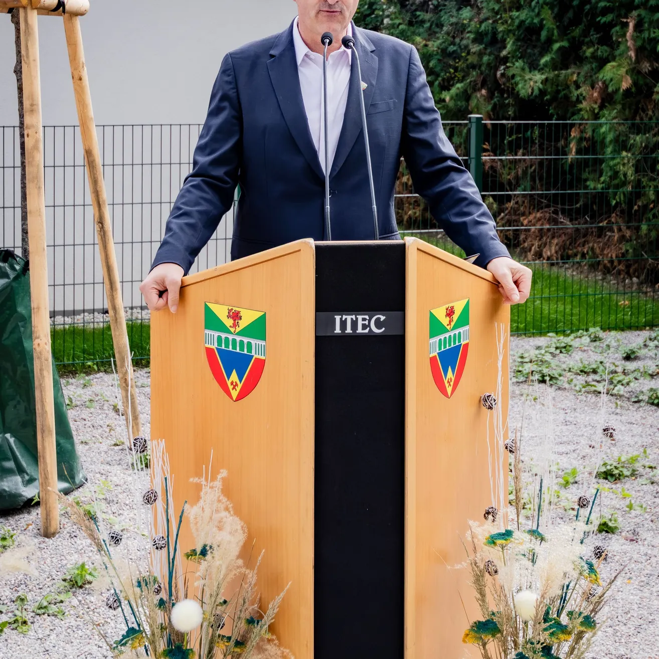 Ein Mann in einem Anzug und mit Brille steht hinter einem Podium mit einem Mikrofon. Das Podium hat ein Logo und Text darauf. Auf beiden Seiten des Podiums befinden sich zwei Vasen mit Blumen.