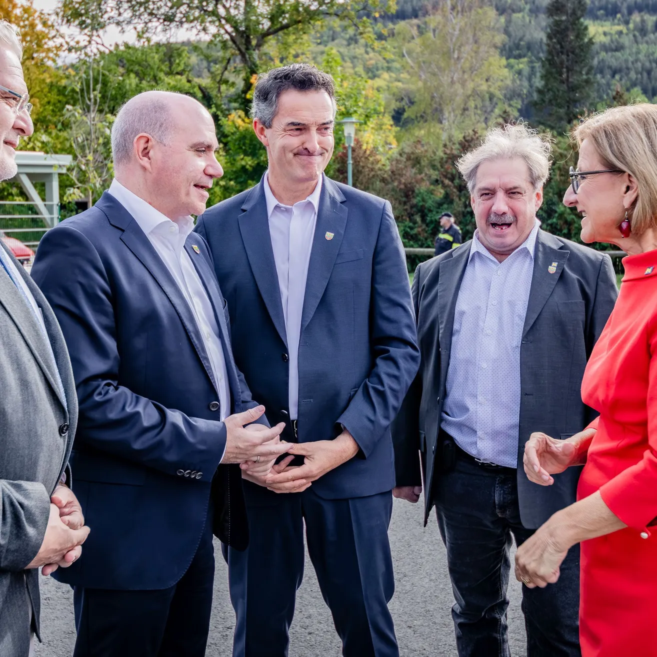 Eine Gruppe von Menschen in formeller Kleidung steht im Freien. Sie alle lächeln und scheinen ein Gespräch zu führen.