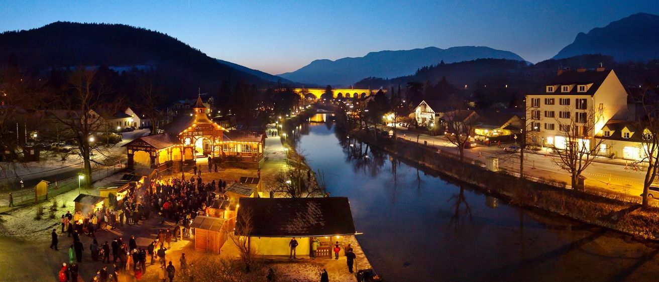 Eine geschäftige Nachtszene einer Stadt am Fluss, mit Menschen, die sich um ein Lagerfeuer versammelt haben, einer Brücke über dem Fluss und Gebäuden, die bei einem blauen Himmel beleuchtet sind.