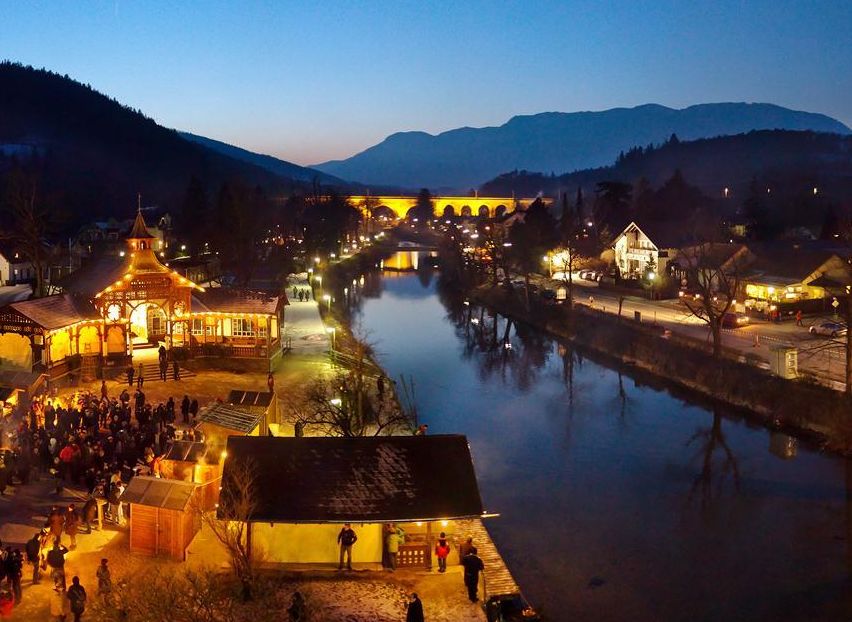 Eine geschäftige Nachtszene einer Stadt am Fluss, mit Menschen, die sich um ein Lagerfeuer versammelt haben, einer Brücke über dem Fluss und Gebäuden, die bei einem blauen Himmel beleuchtet sind.