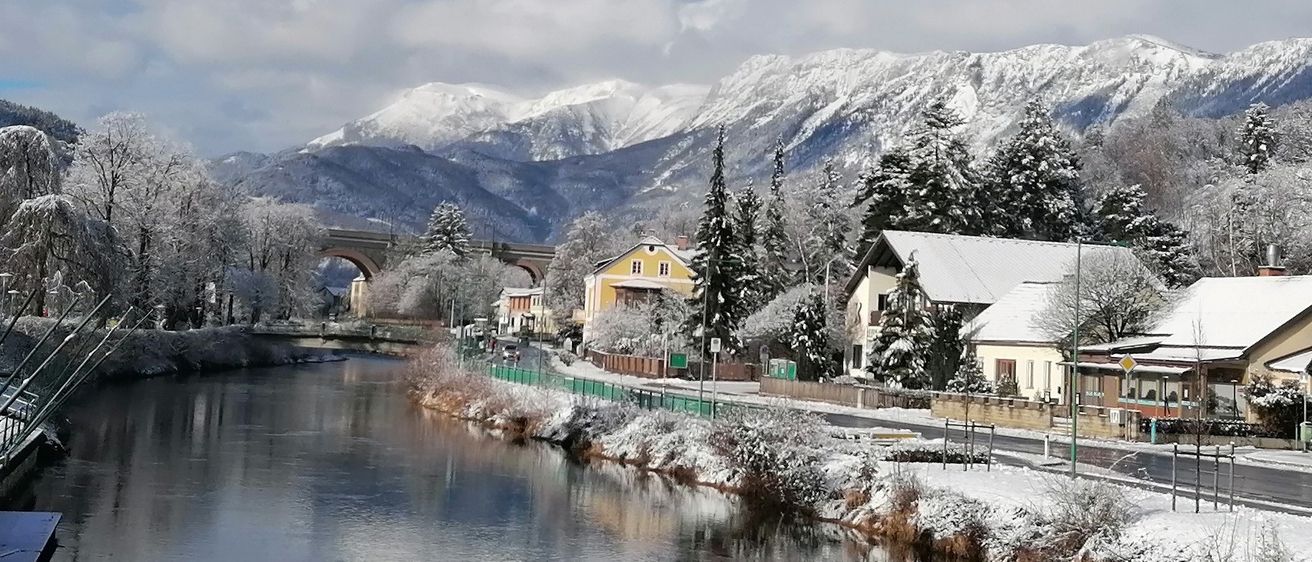 Eine friedliche Winterlandschaft mit einem Fluss, schneebedeckten Bäumen und Häusern vor dem Hintergrund schneebedeckter Berge und einer Brücke unter einem bewölkten Himmel.