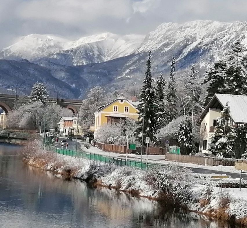 Eine friedliche Winterlandschaft mit einem Fluss, schneebedeckten Bäumen und Häusern vor dem Hintergrund schneebedeckter Berge und einer Brücke unter einem bewölkten Himmel.