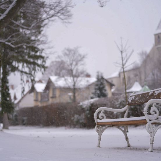 Eine mit Schnee bedeckte Bank steht in einer verschneiten Landschaft mit kahlen Bäumen und einem Gebäude im Hintergrund.